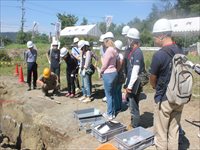 発掘現場でヘルメットを着用した参加者たちが説明を聞きながら作業を見学している写真