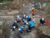 発掘現場でヘルメットを付けた参加者たちが地層を調べている写真