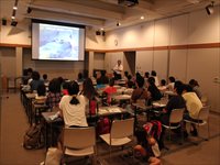 講義室で参加者たちが資料を見ながら講師の解説を聞いている写真