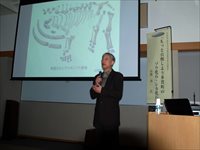 講義室で講師がマイクを持ってスクリーンを使い説明している講演会の写真