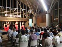 ホールの舞台上で複数の出演者が演奏し客席が鑑賞している様子の写真