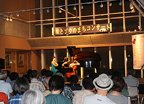 ホールの舞台上で演奏する出演者と客席で鑑賞している来場者の写真