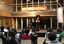 ホールの舞台上でピアノと歌の演奏を行う出演者を客席が鑑賞している写真