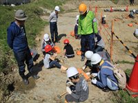 屋外の発掘現場でヘルメットを着けた子供たちと大人が地面を掘って化石発掘体験をしている写真
