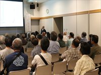 室内の講義室で聴講者たちが椅子に座り前方の講演を聞いている様子を後ろから写した写真