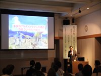 室内の講演会場で登壇者がスライドを使って発表している様子を客席から写した写真