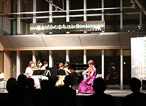 夜の屋内ホールの舞台上でドレスを着た出演者たちが演奏している様子を客席から写した写真