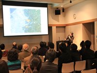 室内の講演会場で登壇者がスクリーンを指し示しながら説明している様子を写した写真