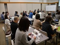 室内の会議室で参加者たちが資料を見ながら講義を受けている様子を後ろから写した写真