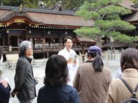 神社の境内でガイド役の職員が参拝者たちに説明をしている様子を後ろから撮った写真