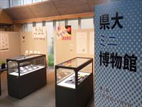 県大ミニ博物館と書かれたパネルとガラスのショーケースに展示品が入っている写真