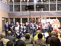 ホールの舞台前でキャラクターと関係者たちが来場者の前に並んで紹介されている写真