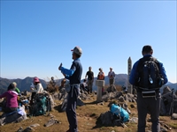 山の山頂付近でガイド役の職員が参加者たちに景色や行程を説明している様子の写真