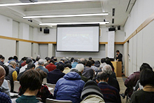 会議室で講師が前に立ち参加者たちがスクリーンを見ながら講演を聞いている写真