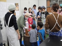 室内でエプロンを着た職員と参加者たちが子どもたちに作業の説明をしている写真