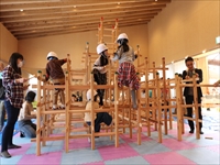 室内の遊び場で木製遊具に登った児童たちを大人たちが見守っている写真