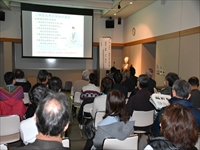 会議室で講師が前に立ち参加者たちがスクリーンを見ながら講演を聞いている写真