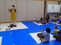 室内でエプロンを着た講師が説明を行い、子供たちが粘土細工に取り組んでいる写真