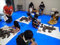 室内で子供たちがブルーシートの上で粘土づくりに集中して作業している写真