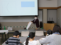 講義室でマスクを着た講師が前に立ち参加者が紙ヒコーキの説明を聞いている写真