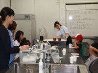 室内の調理室で子供と大人がテーブルを囲み実験や調理をしている様子を横から写した写真