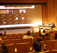 講堂で参加者が手を挙げて質疑をしている講演会を後方から写した写真