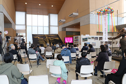 博物館のロビーで来場者が椅子に座って発掘イベントの説明を聞いているのを撮影スタッフが記録している写真