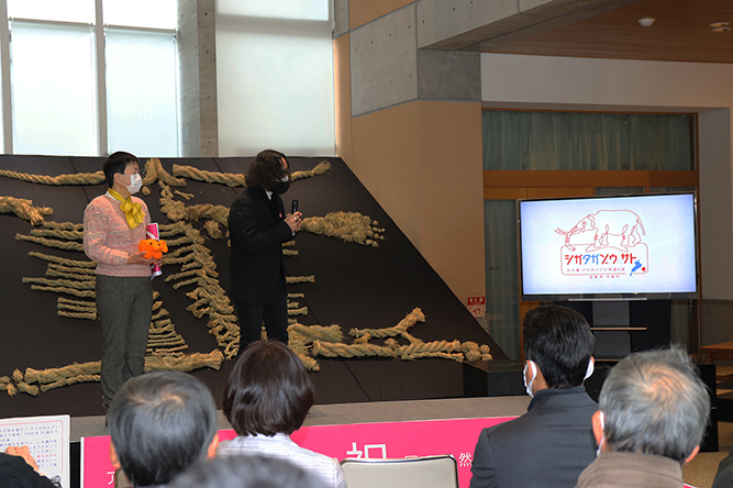 博物館のロビーの特設ステージで登壇者がマイクを持って発掘イベントを紹介しているのを来場者の後ろ姿越しに写した写真