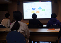 講堂で参加者がスクリーンに映した資料を見ながら講演を聞いている写真