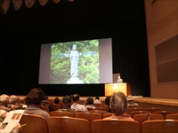 講堂でスクリーンに映し出された映像を見ながら登壇者が発表をしている写真