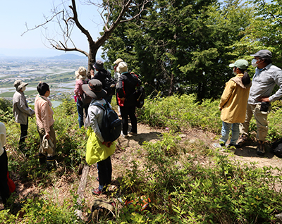 山の展望場所で参加者が景色を見ながら現地解説を聞いている写真