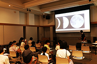 講堂で参加者がスクリーンに映した月の画像を見ながら講演を聞いている写真