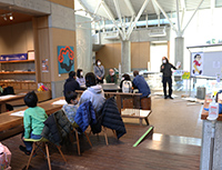 博物館ロビーで来場者が椅子に座り前方の説明を聞いているイベント会場の写真