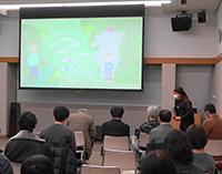 講堂でスクリーンの映像を見ながら来場者が講演を聞いている写真