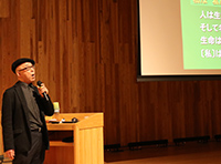 講堂で講師がマイクを持ってステージ横で講演している写真