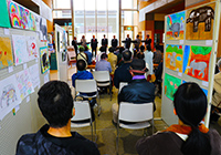 子どもたちが椅子に座って作品展示の通路で発表を聞いているイベントの写真