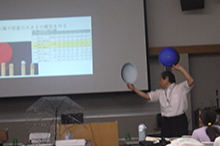 スクリーンの前で白色と青色の球体型の道具を両手に持ち説明をする男性の写真