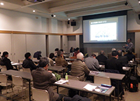 室内で着席しスクリーンを見ながら話を聞く複数人の男女の後ろ姿の写真