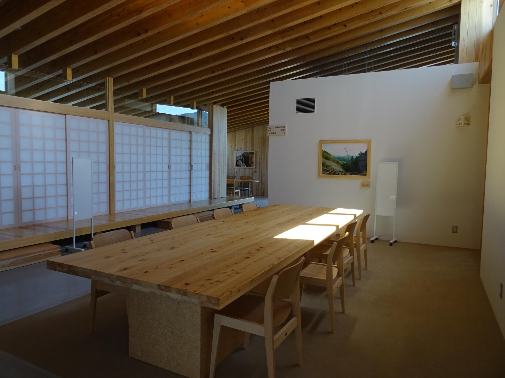 長い木製テーブルと椅子が並ぶつどいの広場を窓側から写した写真