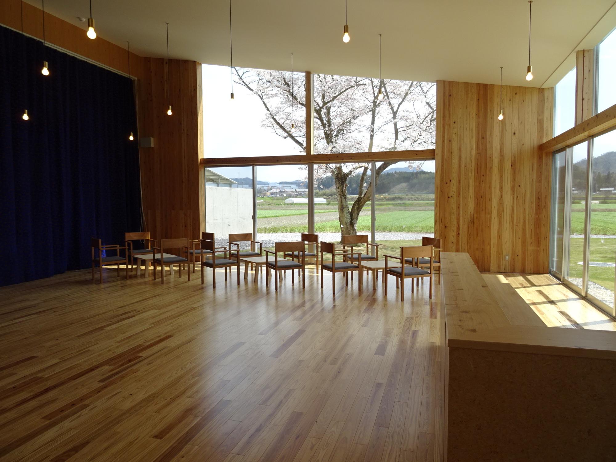 大きな窓から桜と田園風景が見える木造ホールの椅子が並ぶ空間の写真