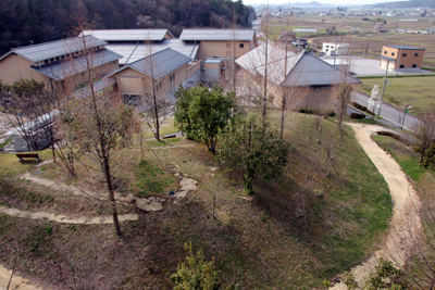 木々が植えられた芝生の丘とカーブした遊歩道、奥に広がる施設群と田園風景写真