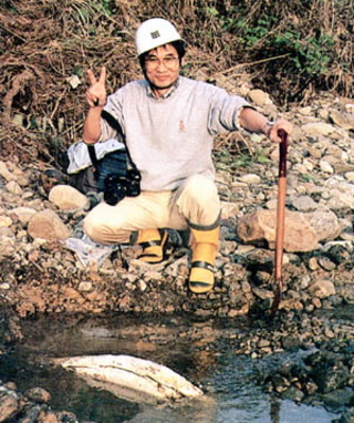 白色のヘルメットを着用し川岸にしゃがみ込みピースサインをしている男性の写真