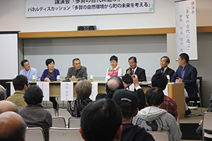 会場前方の壇上でパネリストが着席して討論しているのを参加者が聞いている写真