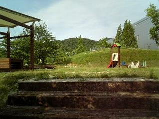 手前には木が立つ芝生の丘と遊歩道があり、奥には大きな屋根の施設が並ぶ風景写真