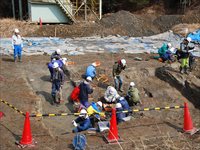 屋外の発掘現場でヘルメットを着用した参加者たちが作業をしている様子を写した写真