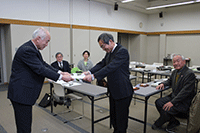 会議室の机の前でスーツ姿の男性2人が表彰状を手渡して授与式をしているのを出席者が見守っている写真