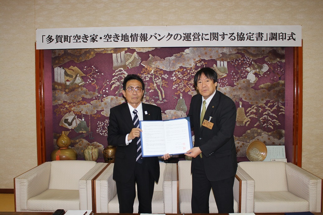 調印式名の書かれた紙が貼られた壁の前で印鑑が押された紙を2人で一緒に見せている写真