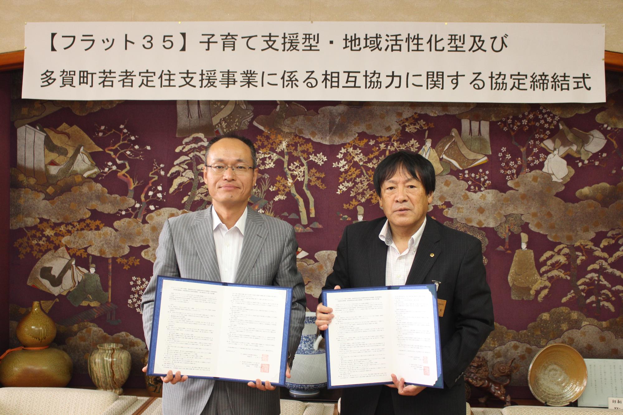 【フラット35】に関する文が書かれた横長の紙を背に印鑑が押された紙を見せているスーツ姿の男性2人の写真