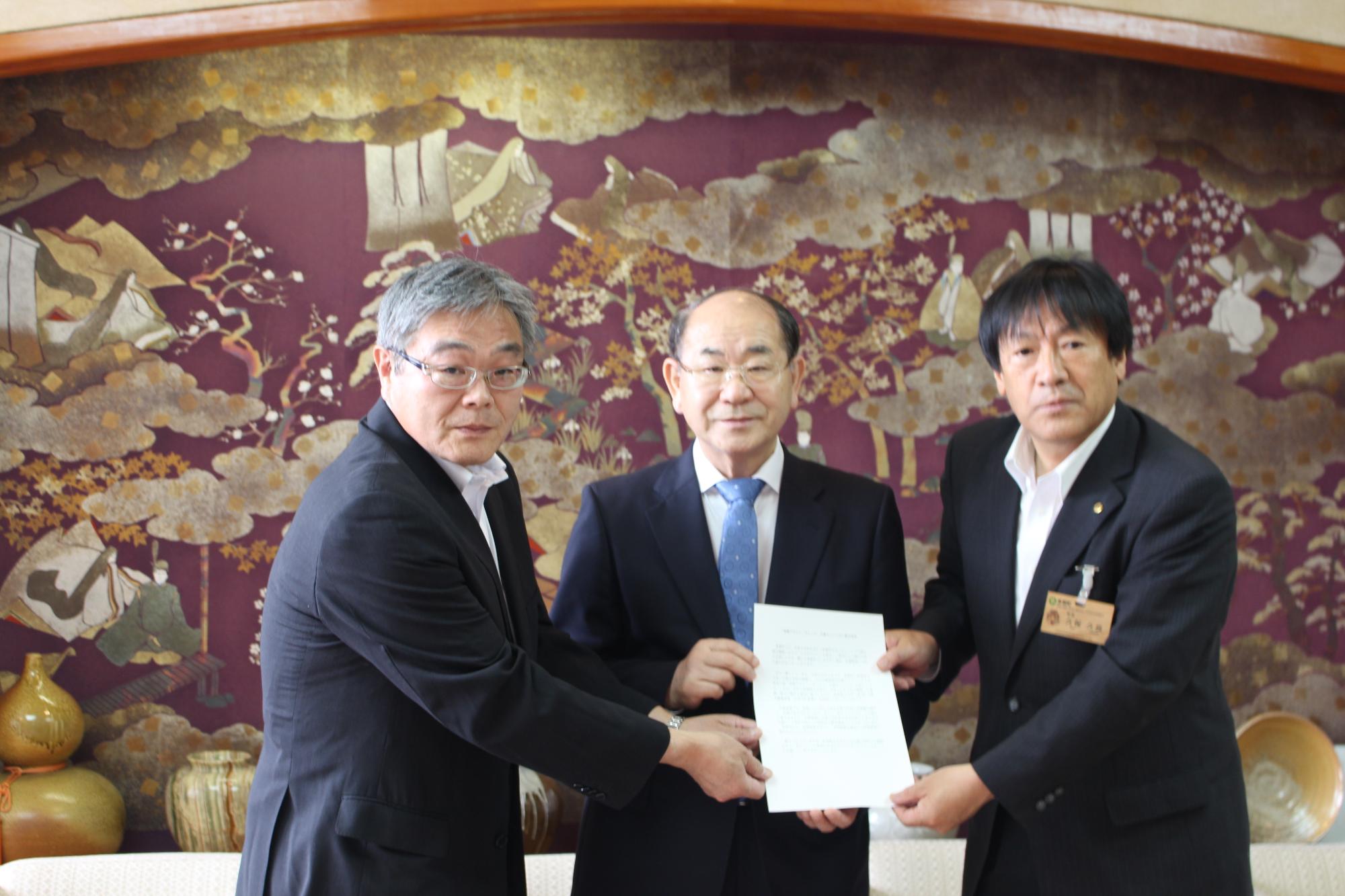 スーツを着た3人の男性が1枚の紙を一緒に持ってカメラに向けて見せている写真