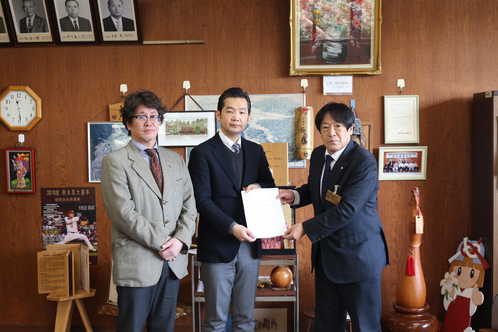 紺のスーツを着た男性二人が一緒に紙を持って見せている横で手を前に組んで立っているスーツ姿の男性の写真